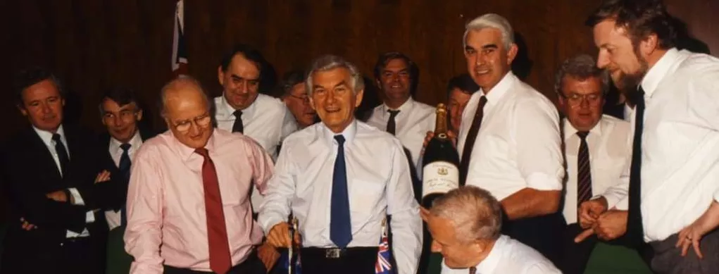 Prime Minister Bob Hawke stands and cuts a square cake, surrounded by a group of men all smiling.