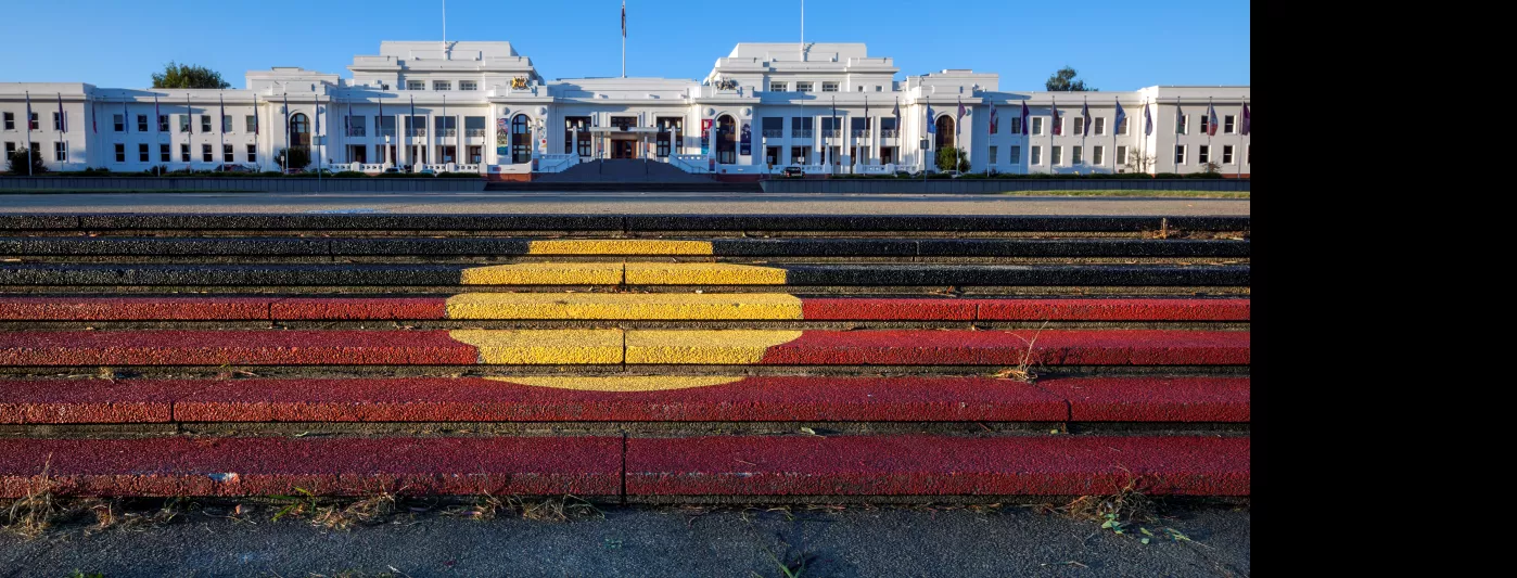 Steps with the Aboriginal flag painted on them lead up to a view of a large long white building, Old Parliament House.