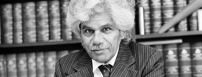 Neville Bonner sits at his desk, wearing a striped suit and tie, a pen in his hand, behind him a full bookshelf.