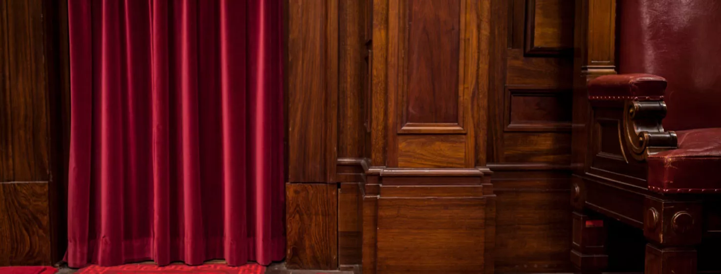 Close up of a red velvet curtin falling to a red carpeted floor with a timber furniture panel next to it.