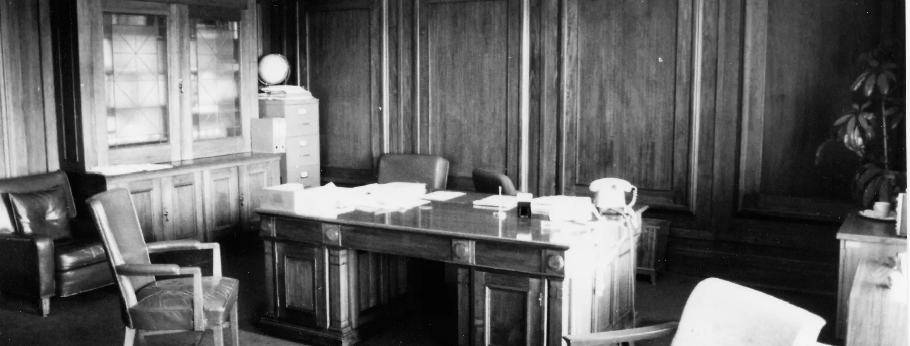 This black and white photograph shows the layout of the Clerk of the Senate’s office in 1985 when it was occupied by Alan Cumming Thom. The timber-panelled room contains a large desk (originally designed by the building’s architect, John Smith Murdoch), glass-fronted built-in bookshelves and a sideboard with an indoor plant. There are a range of visitor chairs and an easy chair in the foreground.