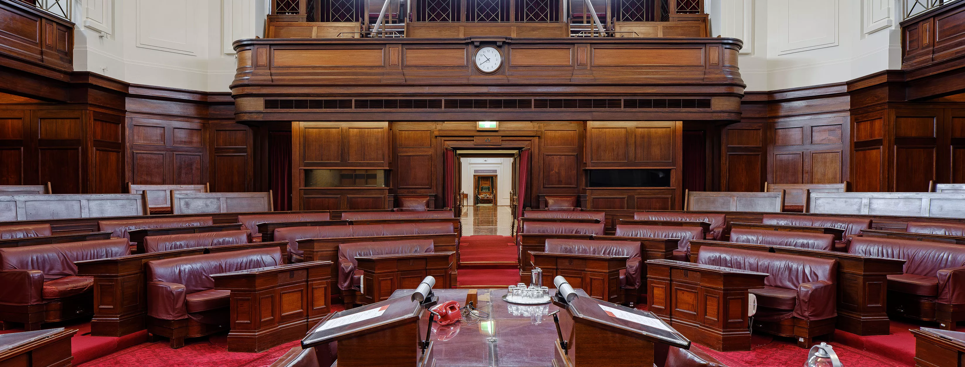 A view out to the Senate chamber. In the foreground are red leather bound books on a long table. Red carpet and red leather chairs in a horseshoe shape with timber panels and art deco lights. 