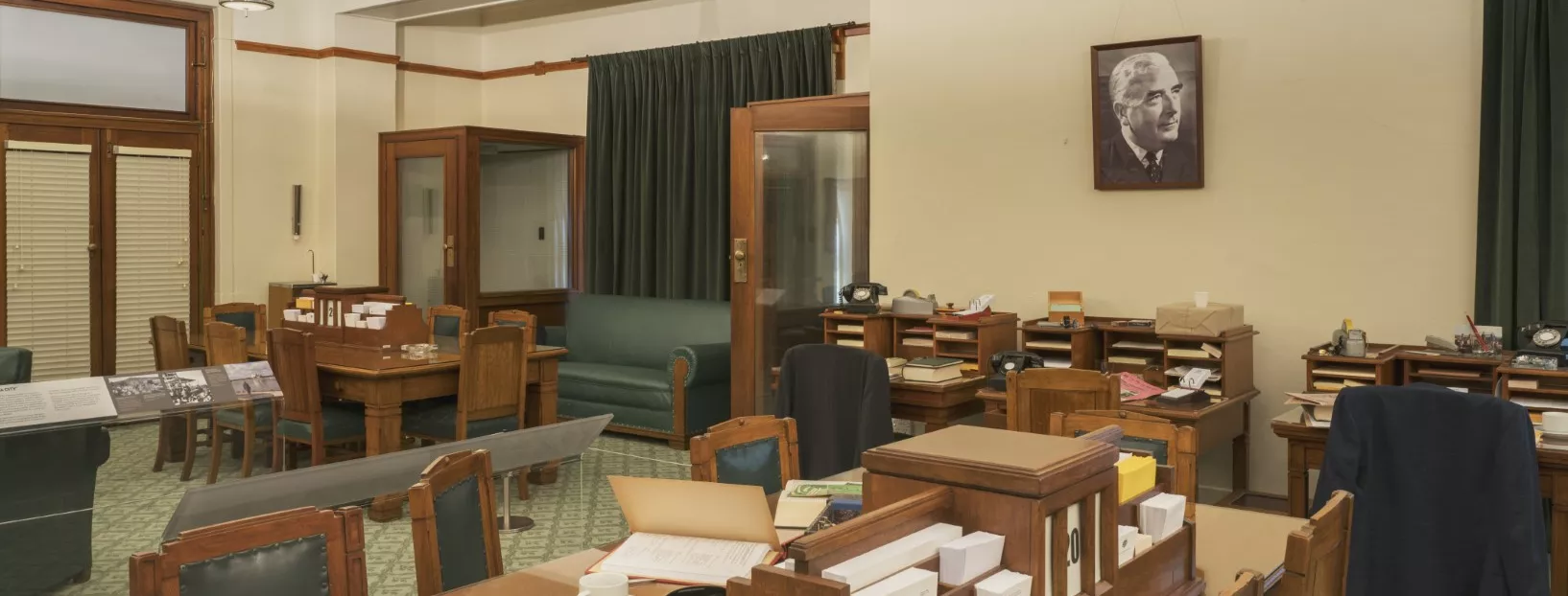 This colour photograph shows the Government Party Room re-created to evoke the atmosphere of 1964. There are two large meeting tables surrounded by chairs, three small desks, and two telephone booths. A large portrait of Prime Minister Robert Menzies is on one wall. The surfaces of the meeting tables and desks are covered with stationery, papers, telephones and ashtrays. 