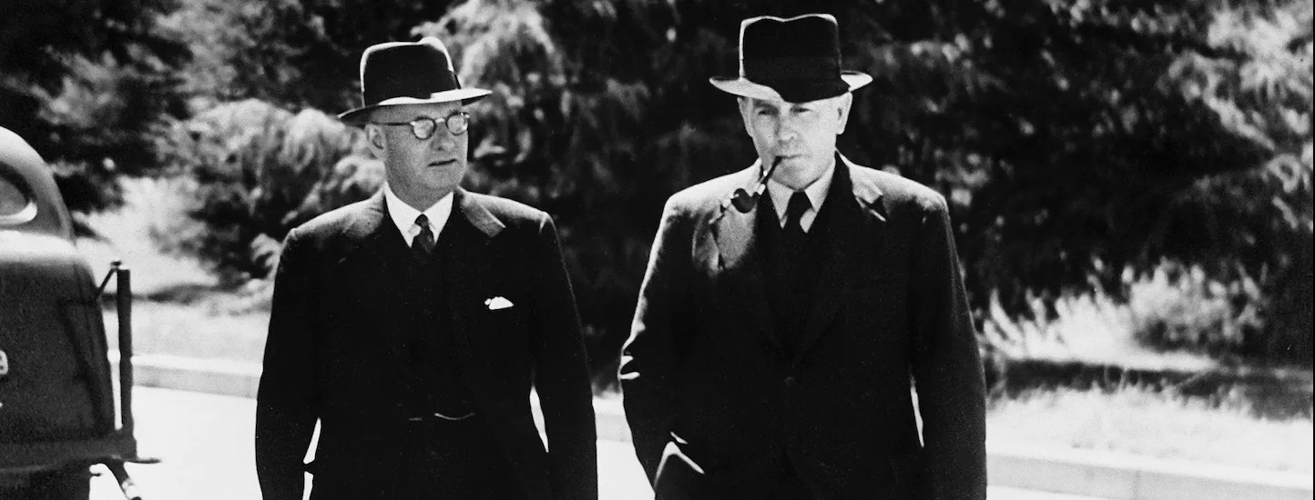 A black and white photo of two men in dark suits, wearing hats, one with glasses and the other with a pipe in his mouth walking. There are two signatures on the photo. One reads John Curtin, the other reads Ben Chifley. 