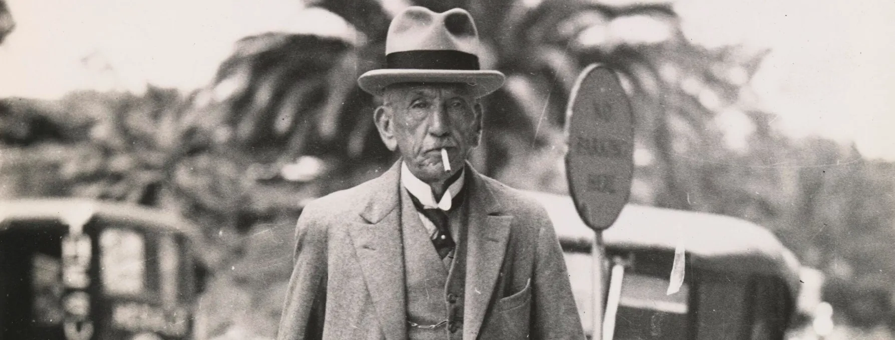 Detail of William Hughes walks down the street wearing a hat and three-piece suit, smoking a cigarette.