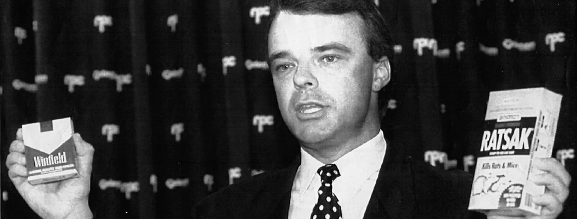 Dr Brendan Nelson, wearing a suit and spotted tie, holding a 'Winfield' cigarette box in one hand and a 'Ratsak' box in the other.