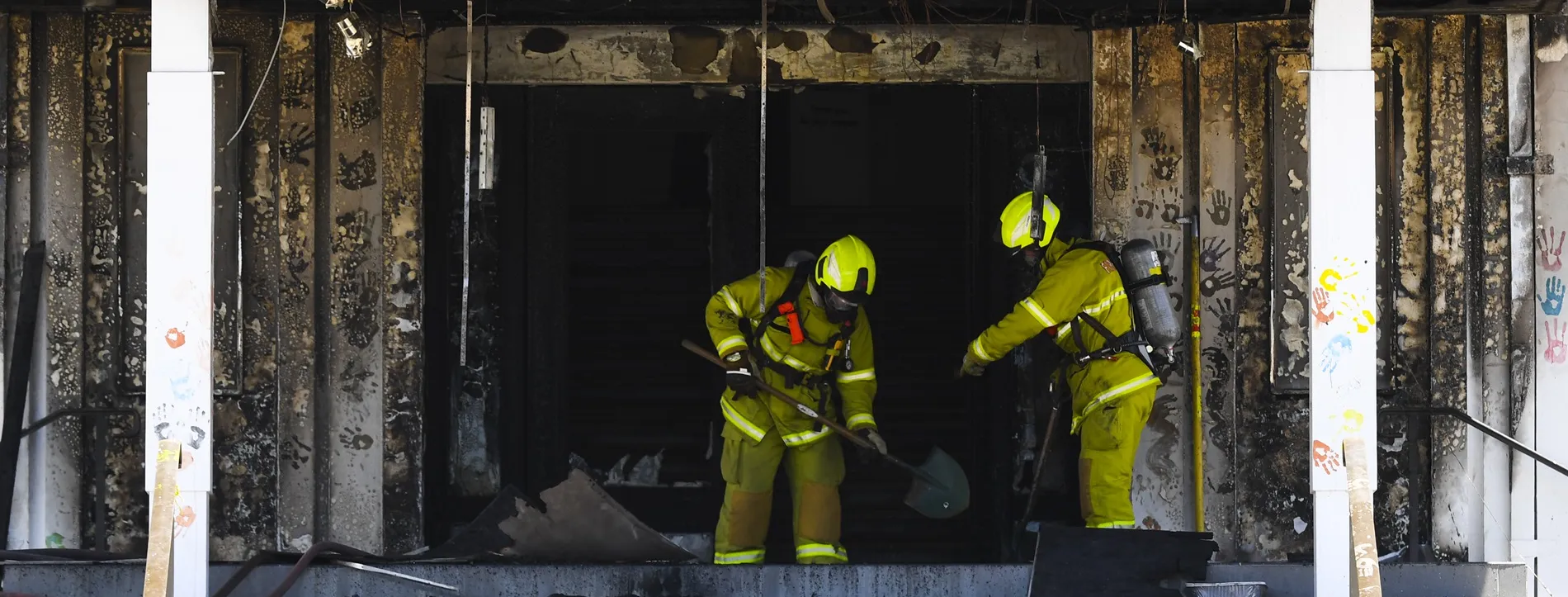 Two firefighters dressed in yellow hi-vis clothing and protective equipment, hold shovels and assess the fire damage to the front entrance of Old Parliament House.