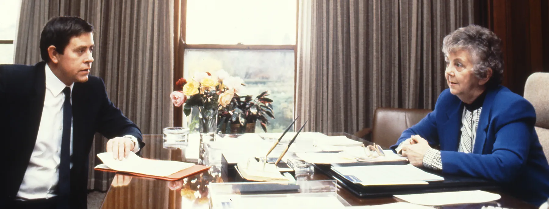 This professional colour photograph shows Principal Private Secretary John Porter and Speaker Joan Child sitting across from each other at the Speaker’s desk. John is wearing a dark suit, white shirt and dark tie and Joan is in a white blouse and vibrant blue jacket. They are discussing the daily program for the House of Representatives Chamber. There is a vase of roses, stationery and paperwork on the desk. In the background the curtains have been drawn to reveal a large east-facing window.