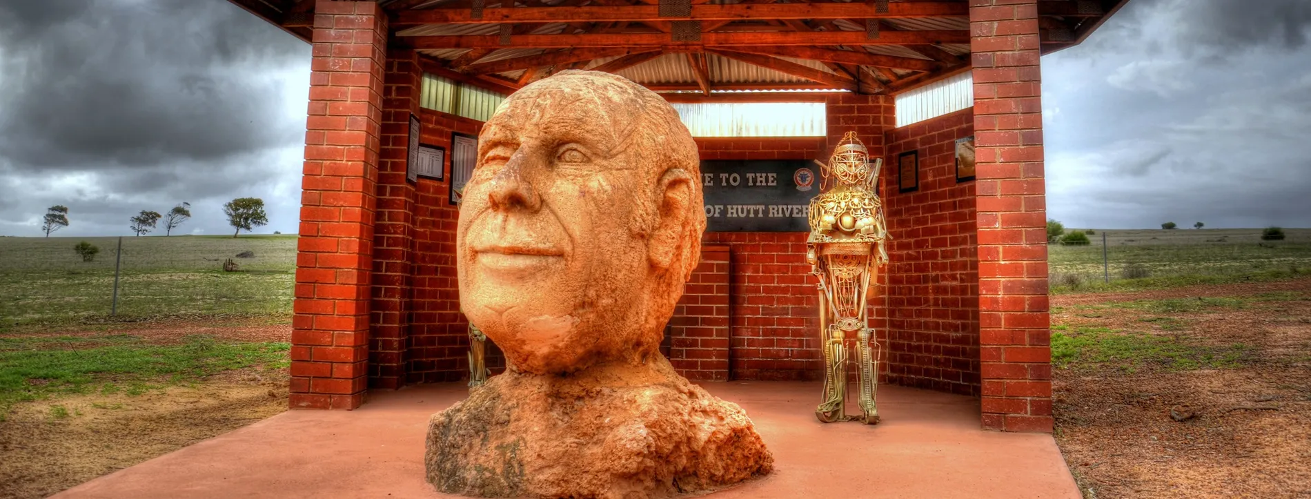 A hand-carved rock bust depicting the head and shoulders of 'Prince' Leonard George Casley, positioned in front of a redbrick shelter in a paddock.