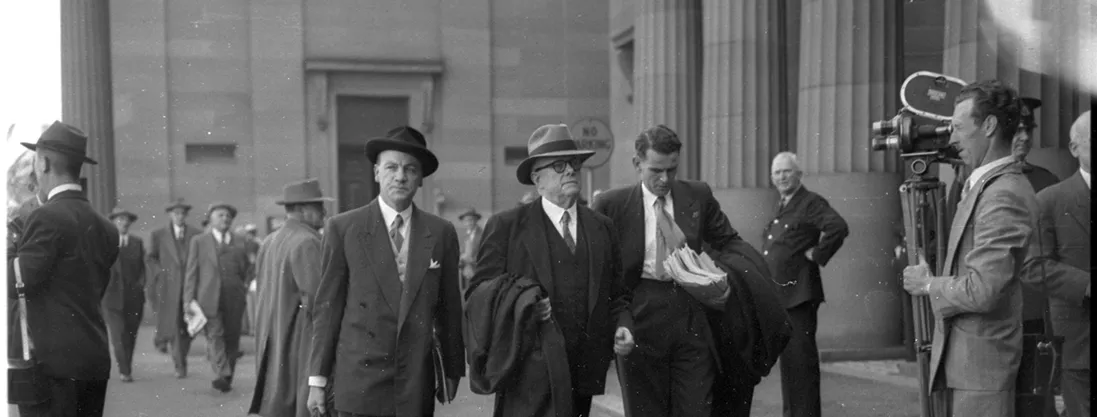 Dr Evatt is flanked by two assistants as he enters the Petrov Royal Commission in Darlinghurst. Men are walking nearby, with a man standing with a camera recording them.