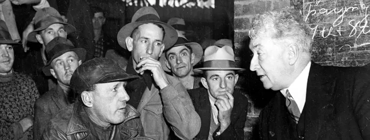 Prime Minister Lyons leans across a desk to chat to wharf workers. A briefcase with the initial A.S. is on the desk. 