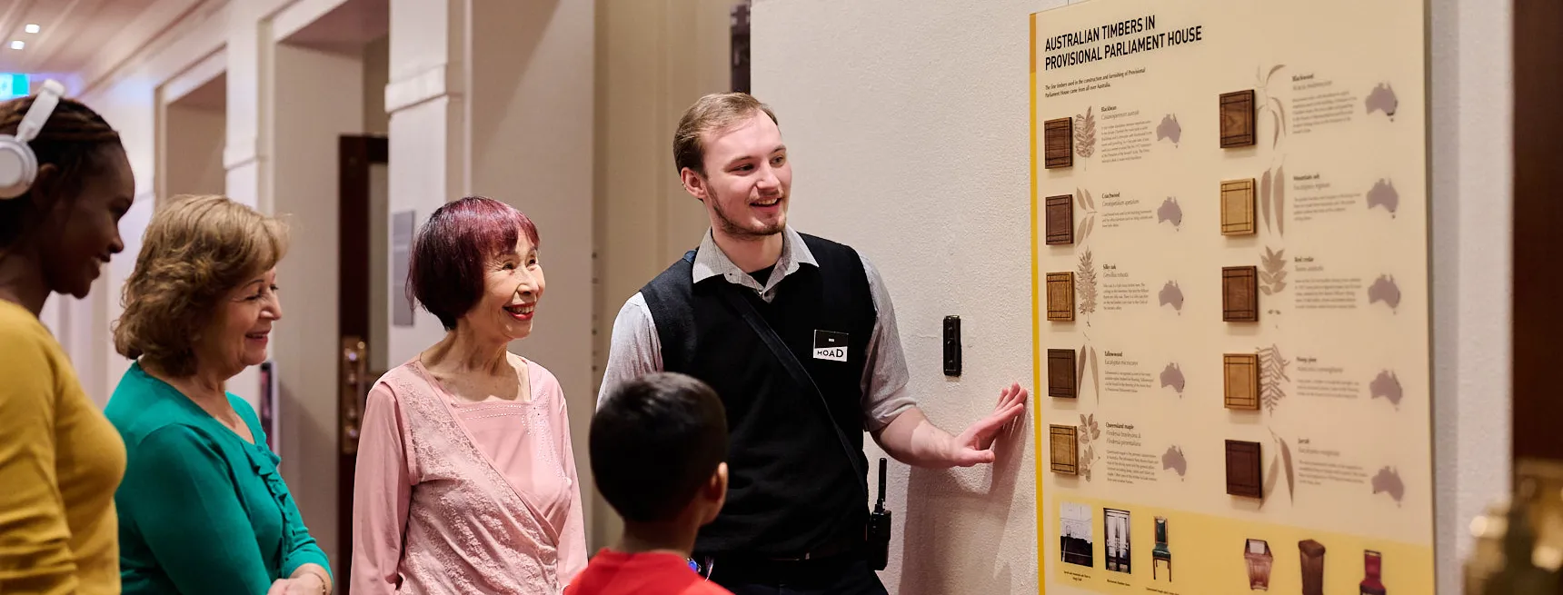 A Museum staff member and a group of people in brightly coloured tops stand in a hallway with red carpet looking at a board displaying Australian timbers used in Old Parliament House.