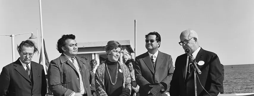 Reg Withers stands behind an Australian flag at a microphone. There are four people to his left. Water is in the background.