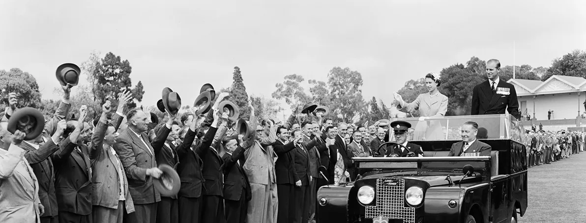 The Queen and Prince Phillip are standing in the back of a car waving to a crowd who are all waving their hats to them.