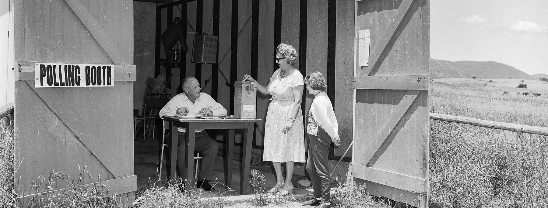 A woman puts a voting card in a ballot box at a shed in a paddock being used as a polling booth. A man sits at a table and a child looks on. 