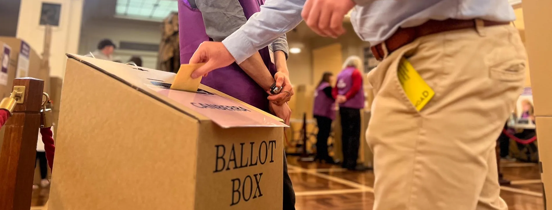 A voter puts a ballot paper in a cardboard ballot box while an AEC official dressed in a purple AEC vest looks on. They are in King's Hall in Old Parliament House.