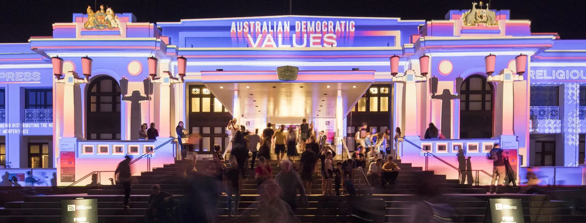 The front of Old Parliament House at night lit with multi-coloured lights and the words 'Australian democratic values'. A crowd of people walk up the stairs into the building.