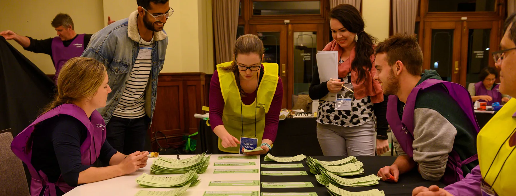 Officials in yellow and purple vests count green ballot paper votes on a table in Old Parliament House. Officials in yellow and purple vests count green ballot paper votes on a table in Old Parliament House.