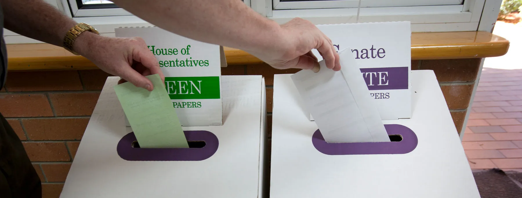 Hands place green and white ballot papers into white and purple ballot boxes.