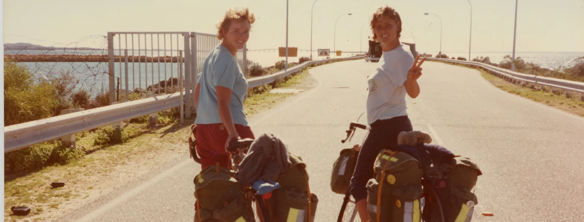 Two women on bikes on a road near the ocean.