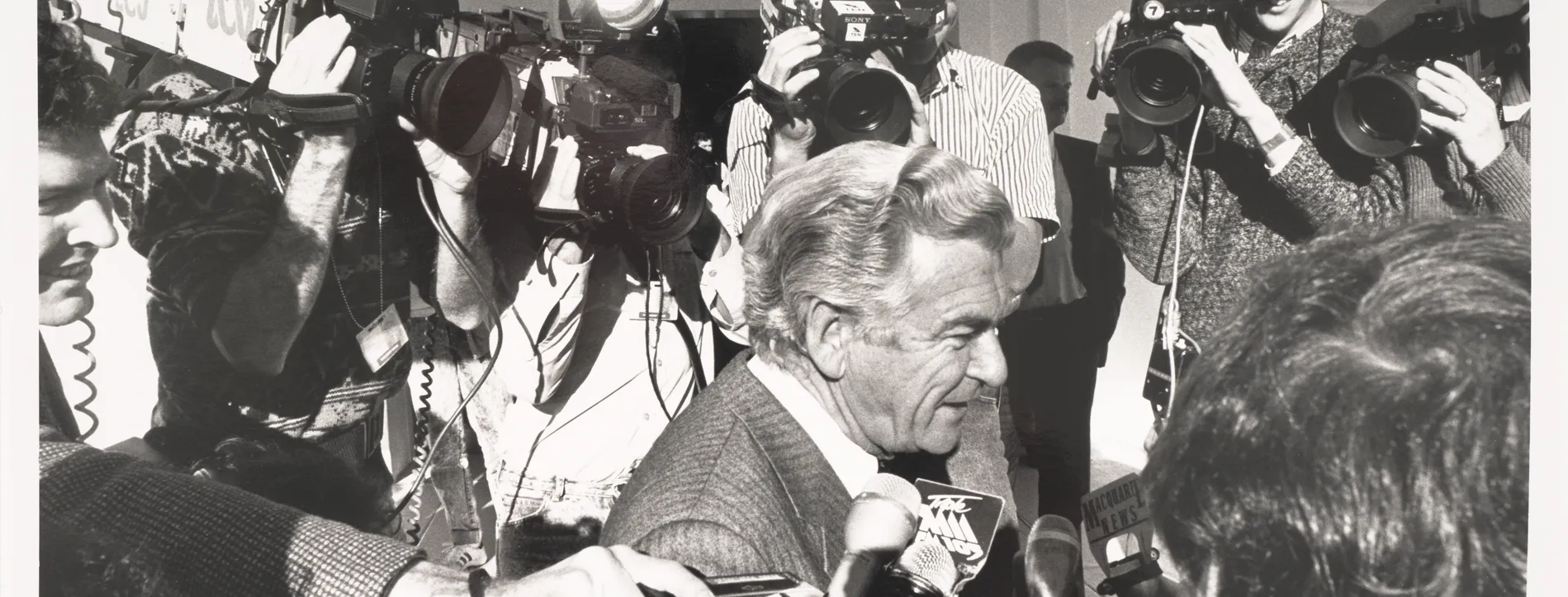 Prime Minister Bob Hawke arriving at Parliament House being swarmed by the media, all pointing cameras in his face.