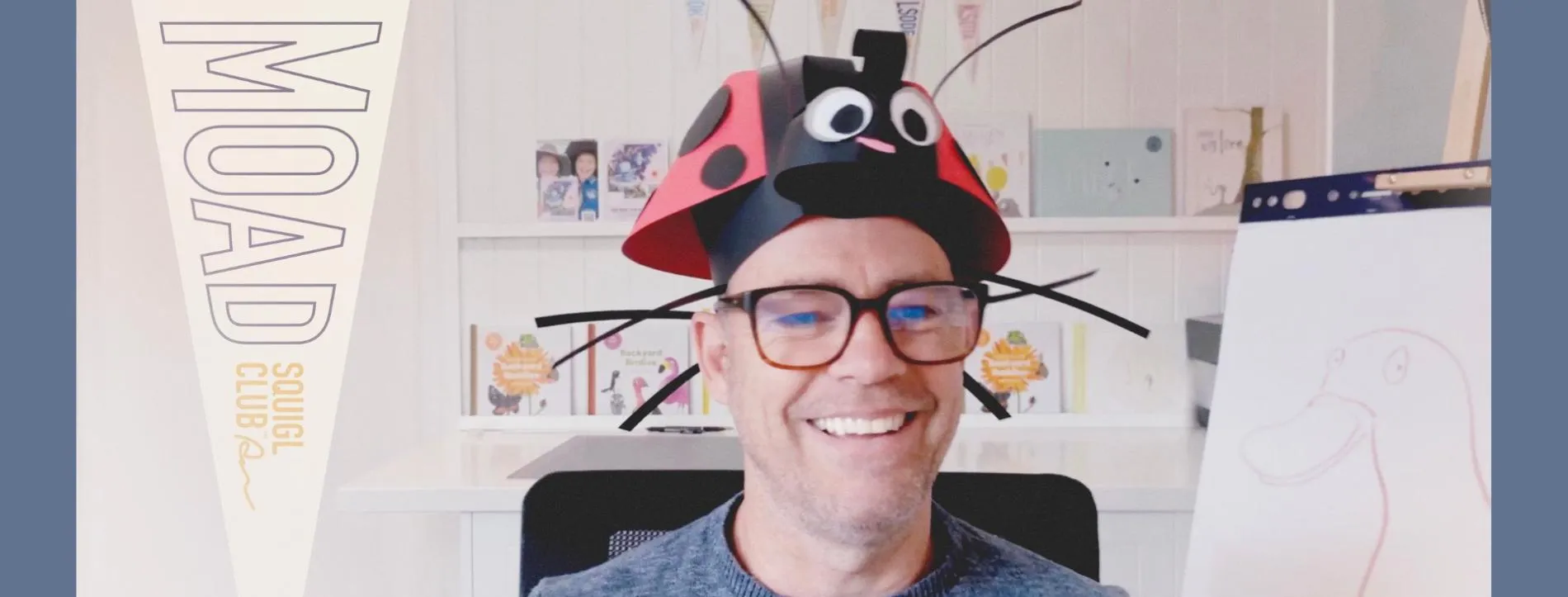 A man wearing a ladybug hat sits in front of a bookcase of children's books.