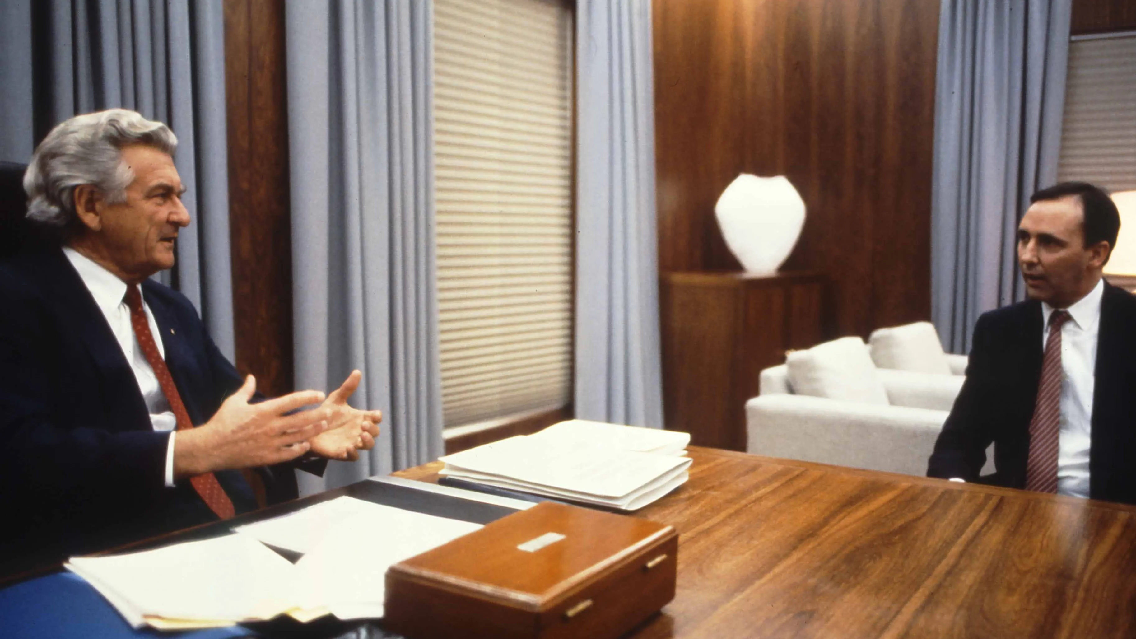 This colour photograph captures a conversation between Prime Minister Bob Hawke seated on the left and Treasurer Paul Keating seated on the right. The photo shows the corner of the desk with Hawke sitting behind the desk while Keating sits to the side with both men facing each other. Both are dressed in black suits with white shirts and red ties contrasting the pale blue curtains in the background.  