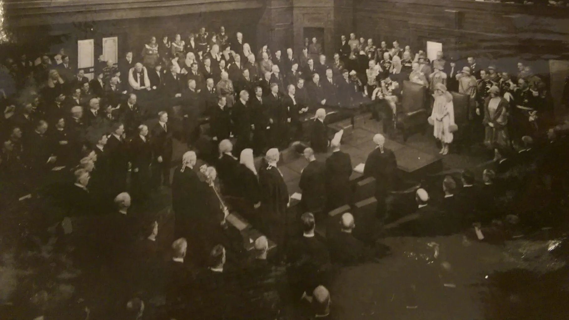 This black and white photograph shows the Duke of York reading a message from his father the King in the Senate Chamber. The room is crowded with at least 200 people, mostly men with some women all wearing their best. Men are dressed in suits, military uniform or gowns and wigs while the women wear formal dresses with hats and gloves. The crowd is in a horseshoe shape around the central table while the Duke stands at the head of the table.