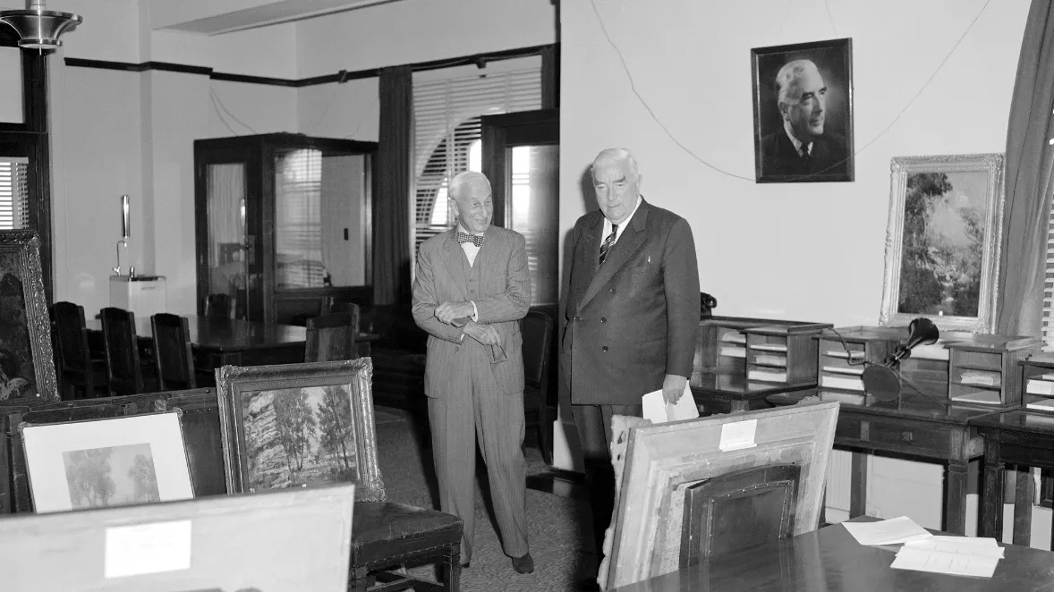 This black and white photograph shows two suited men standing side by side in the Government Party Room in 1962. On the left is Sydney businessman and philanthropist SH Ervin with a jaunty bow tie, on the right a more conservatively dressed Prime Minister Bob Menzies. They are surrounded by Australian paintings which are leaning against chairs, tables and walls. The two men are viewing the paintings that Ervin was donating to the National Collection. In the background are meeting tables and telephone booths