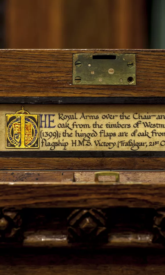 Cursive script on a label on the side of the Speaker's Chair with the words 'The Royal Arms over the chair are carved in oak from the timbersof Westminster Hall (1399); the hinged flaps are of oak from Nelson's flagships HMS Victory (Trafalger, 21st October, 1805).