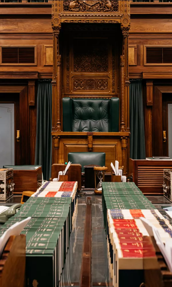 The table of the House of Representatives chamber looking up to the Speaker's Chair at the end. Papers and books cover the tables. The chair is wooden, leather and ornate.