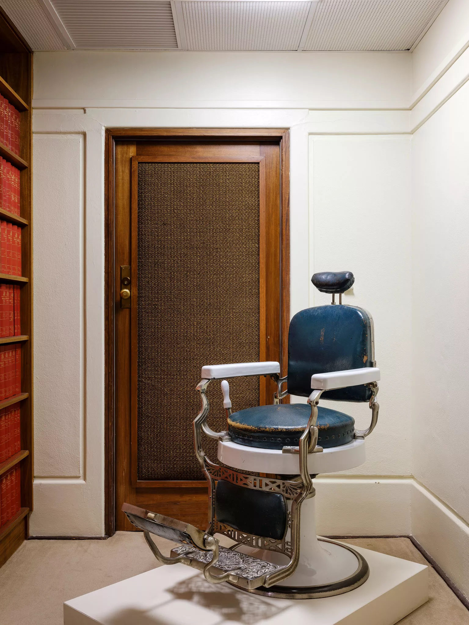 A blue leather barber's chair with white handles displayed in a room with a brown wooden door behind it. 