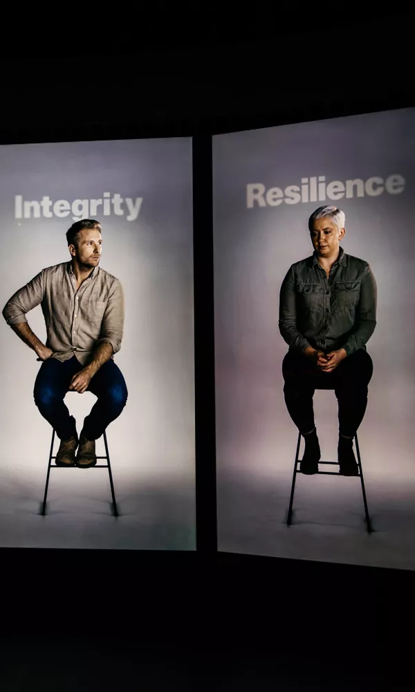 Four video screens with a person sitting on a stool on each screen. The words integrity, resilience and duty appear above them. Four video screens with a person sitting on a stool on each screen. The words integrity, resilience and duty appear above them.