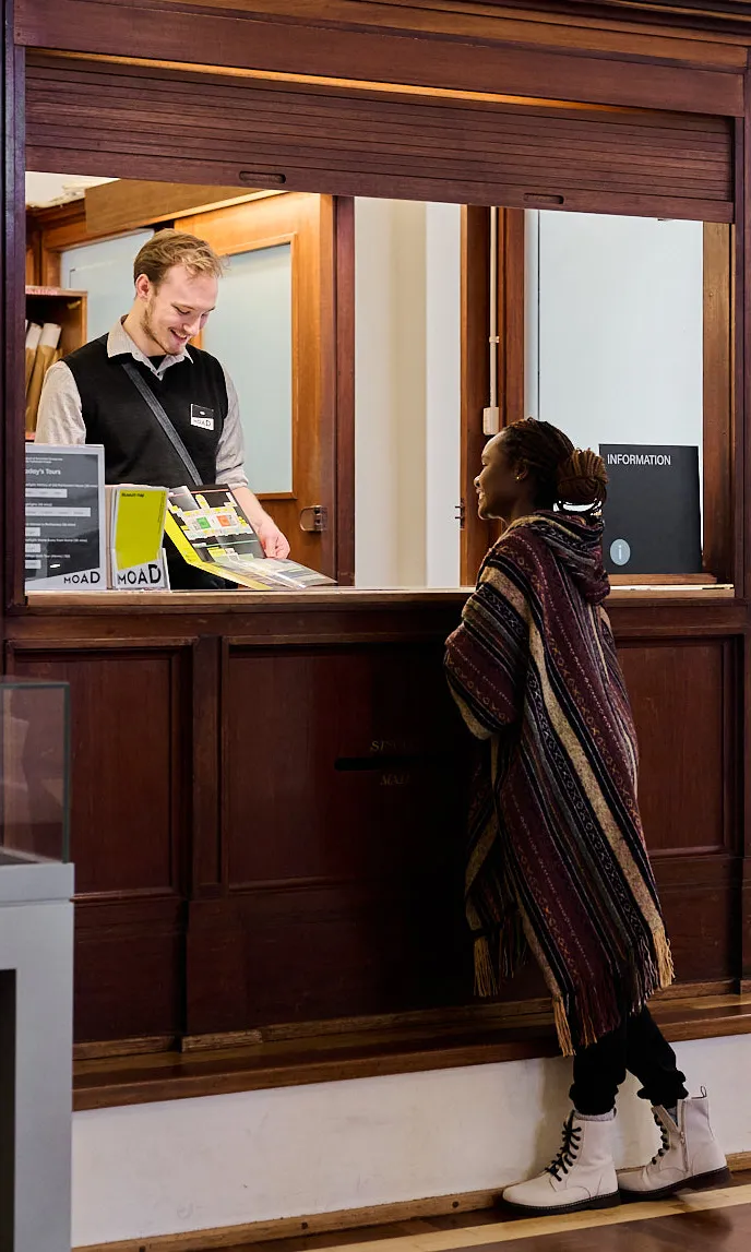 A Museum Experience Officer wearing a black vest and name tag standing inside an information booth at MoAD at Old Parliament House assists a woman with a brochure about the Museum.