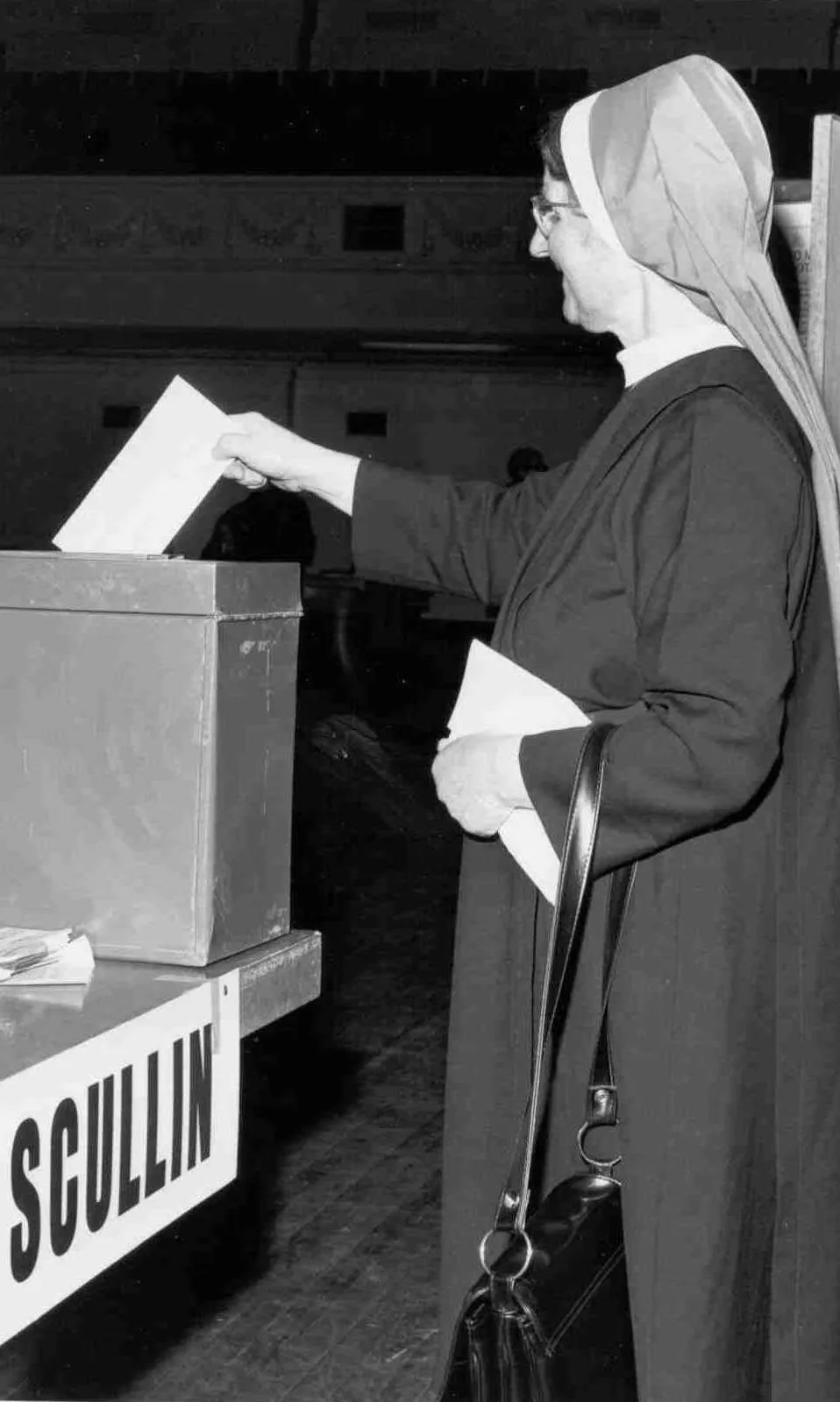 A nun wearing a habit puts a ballot paper into a ballot box marked 'Scullin'.