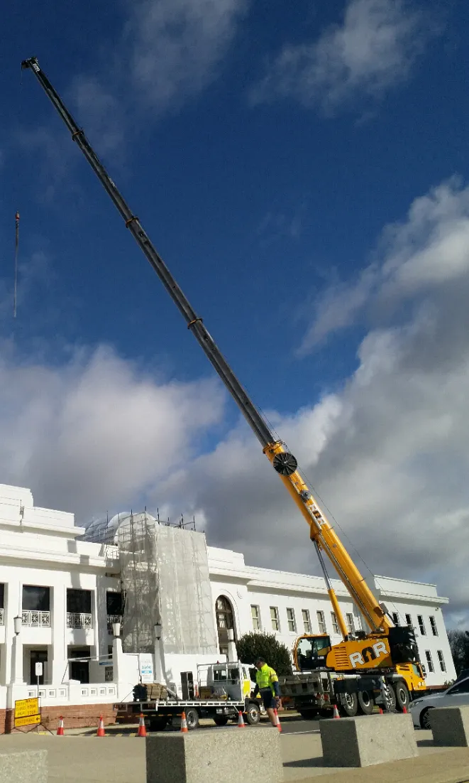 A large yellow crane sits in front of the white front exterior of Old Parliament House. A man in a yellow hi-vis jacket stands next to it.