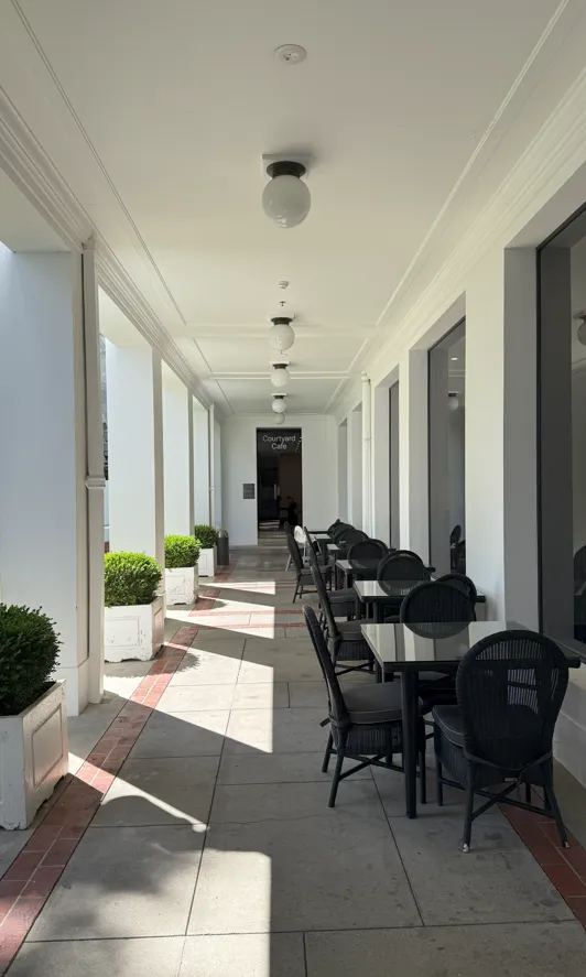 A walk way with a white ceiling and a green courtyard on the left. Black wicker tables line the path to a door. 