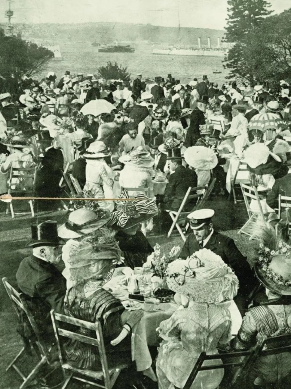 A large group of people enjoying tea at garden tables on the lawn, boats passing on the harbour in the background.