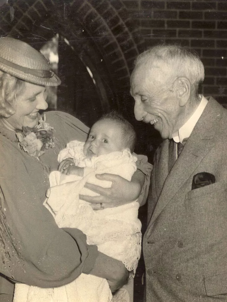 Billy Hughes wearing a coat and standing with a woman holding a baby.