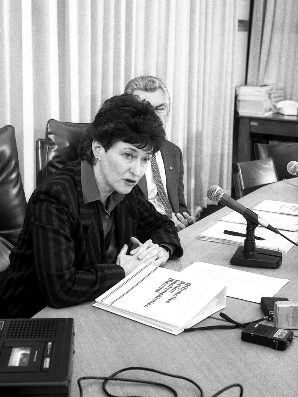 Senator Susan Ryan sit at a table, next to Prime Minister Bob Hawke, speaking into a microphone with a binder in front of her labelled 'Affirmative action implementation manual'.