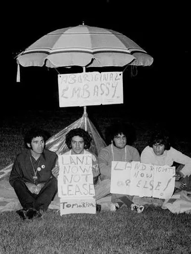 Four men sit on the ground under a beach umbrella with signs that read, 'Aboriginal Embassy', 'Land now not lease tomorrow' and 'Land rights now or else!'.