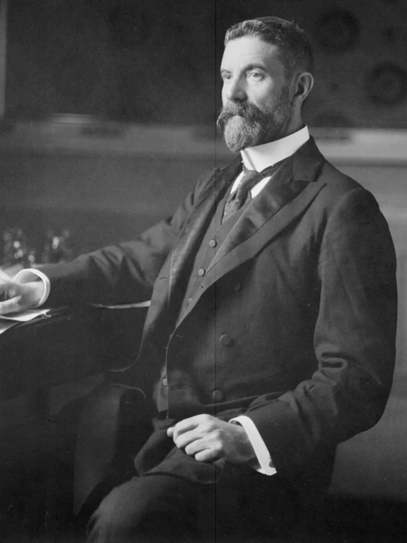 Alfred Deakin c.1901. Photograph National Library of Australia Alfred Deakin, wearing a three-piece suit and seated at a desk covered in paper documents.