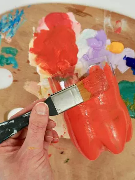 A close up of a hand holding a paint brush painting a plastic bottle red. A close up of a hand holding a paint brush painting a plastic bottle red.