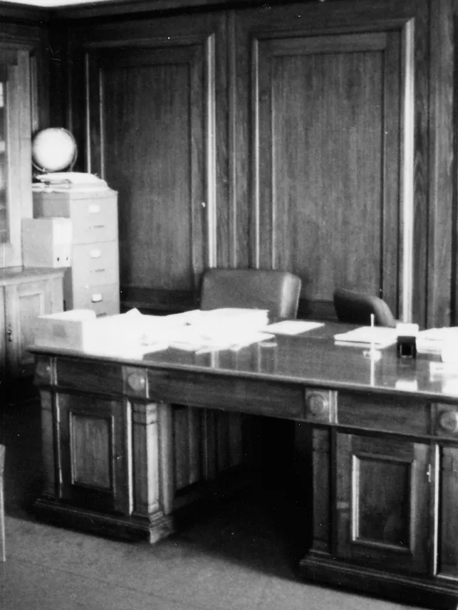 This black and white photograph shows the layout of the Clerk of the Senate’s office in 1985 when it was occupied by Alan Cumming Thom. The timber-panelled room contains a large desk (originally designed by the building’s architect, John Smith Murdoch), glass-fronted built-in bookshelves and a sideboard with an indoor plant. There are a range of visitor chairs and an easy chair in the foreground.