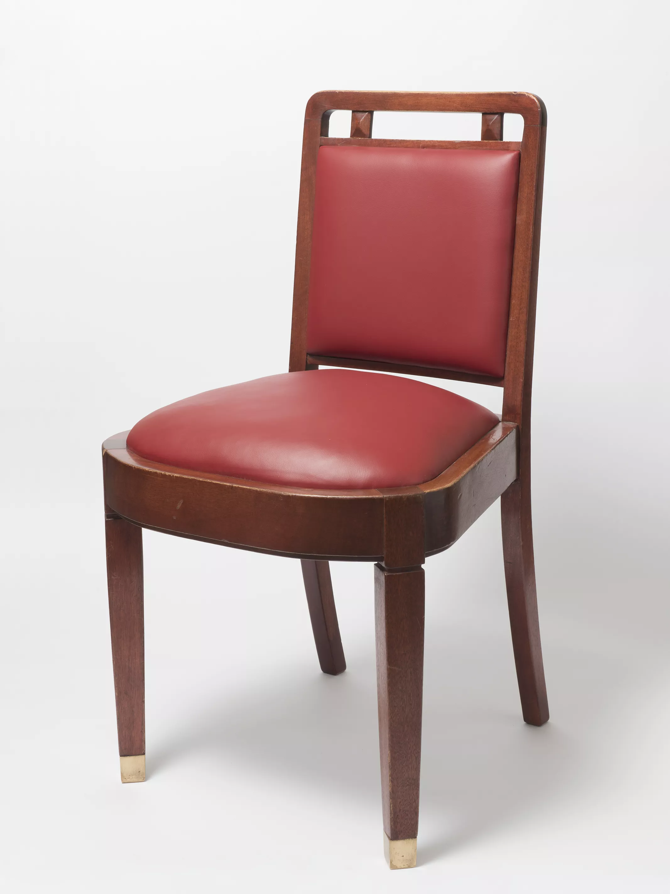 A wooden chair with cherry red leather seat cushion and back, and square legs with protective brass caps on the base of each leg. 