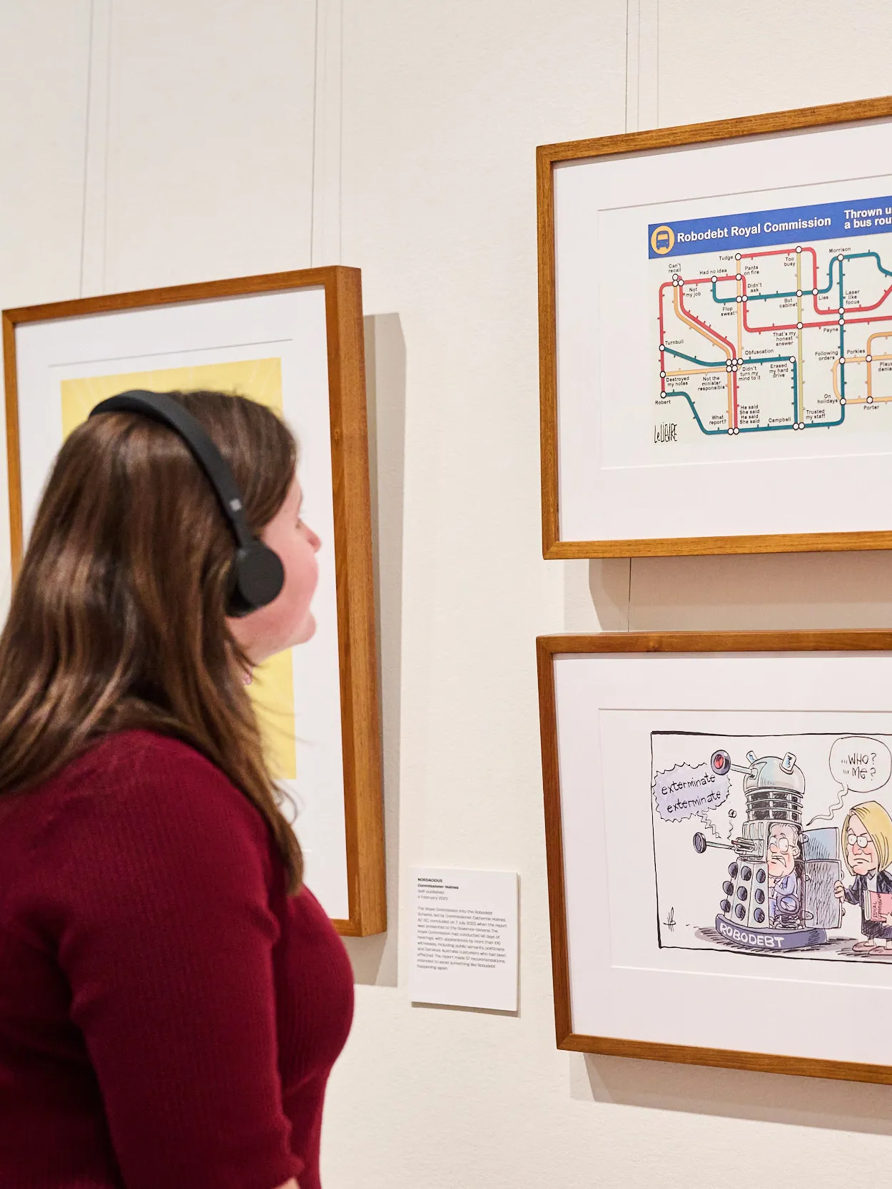 A woman wearing a dark red top and black headphones looks at framed cartoons with a caption featuring the Audio Description logo. A woman wearing a dark red top and black headphones looks at framed cartoons with a caption featuring the Audio Description logo.