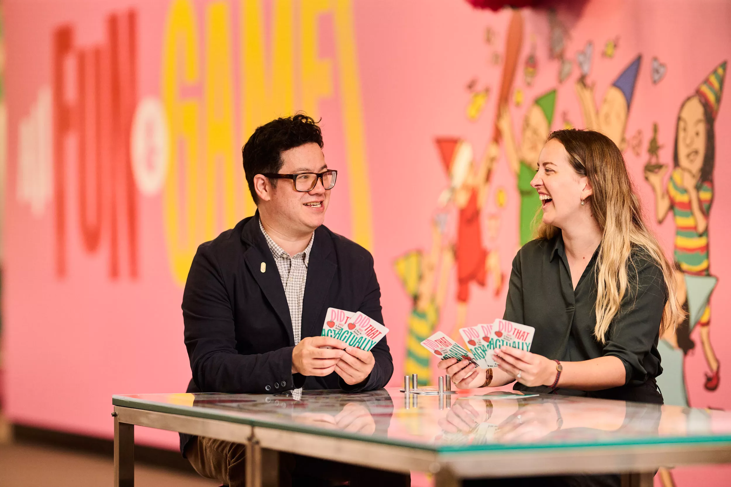 Two people seated at a table playing a card game in front of a brightly decorated wall. Two people seated at a table playing a card game in front of a brightly decorated wall.