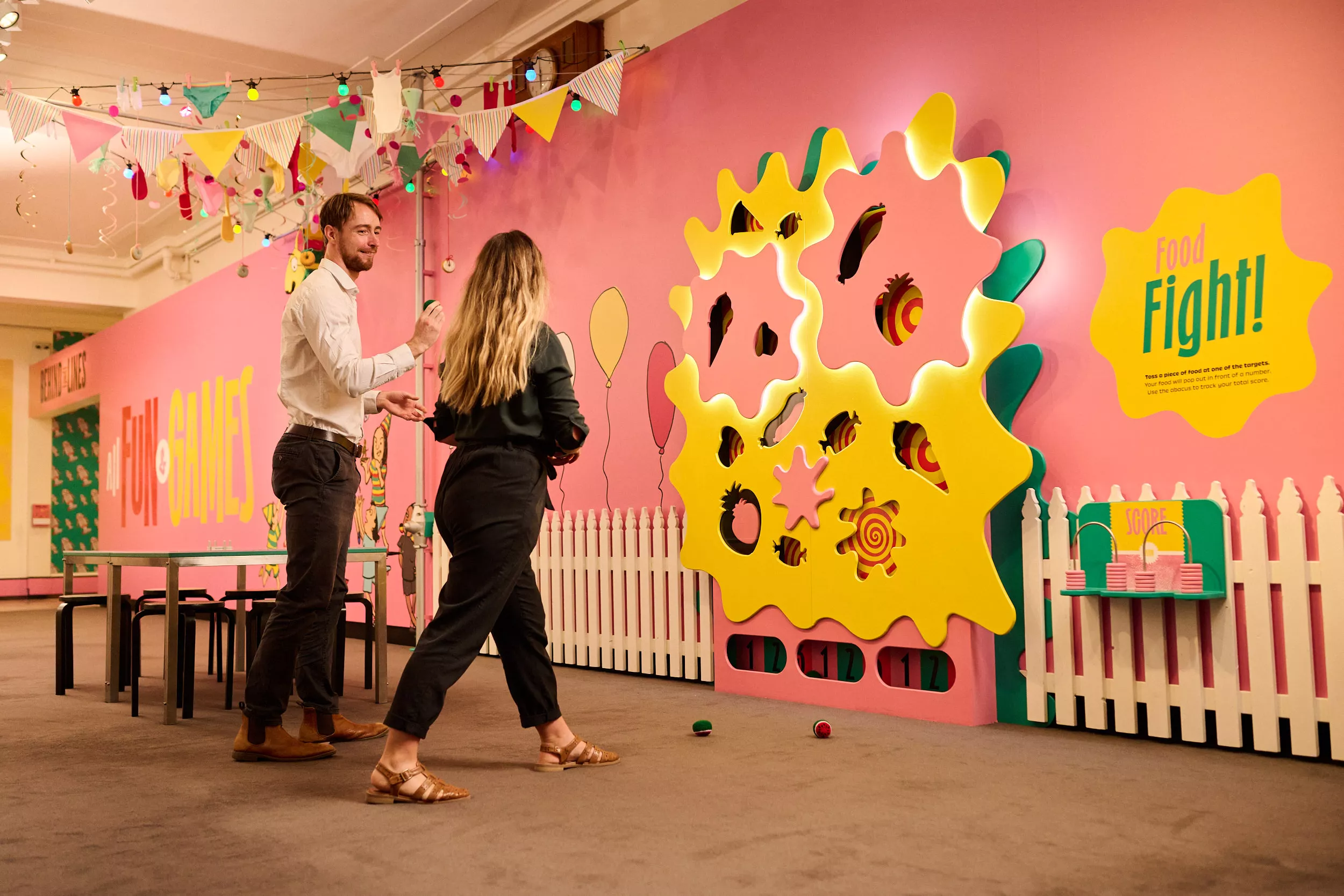 Two people play a game throwing balls into fruit-shaped holes on the wall.