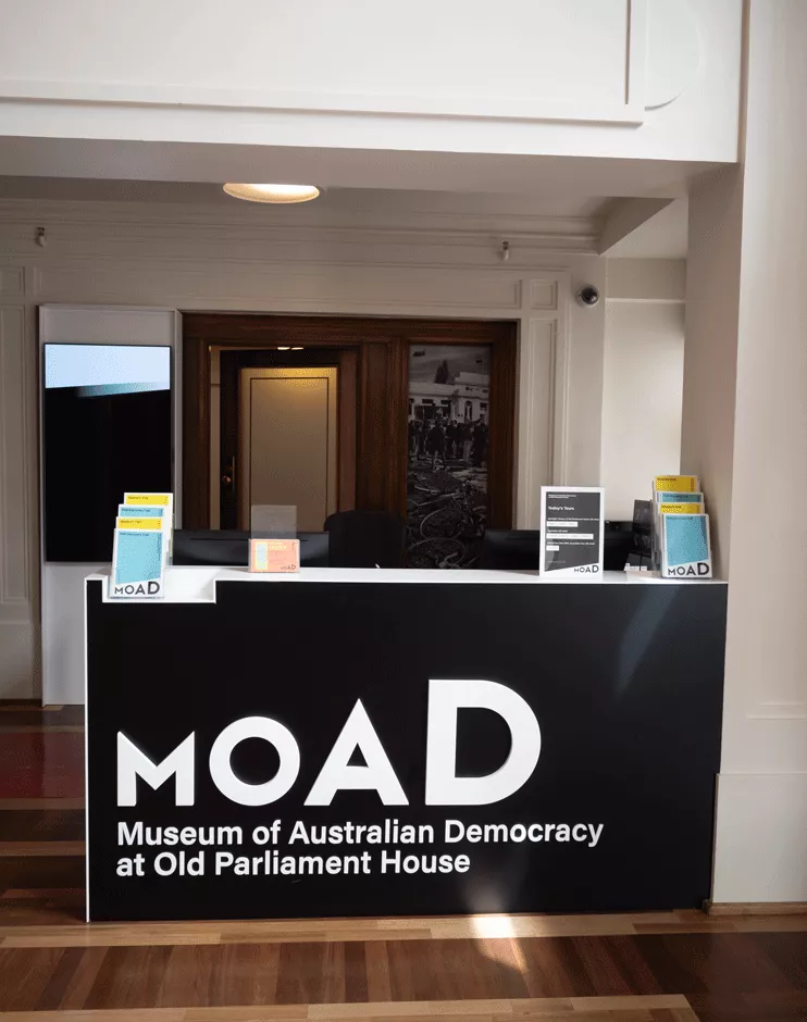 A front desk with pamphlets on top of it and a large black sign with the words 'MoAD Museum of Australian Democracy at Old Parliament House.'