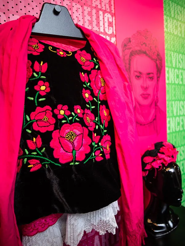 A bright pink coat over a black top with embroidered pink flowers hangs on a pink wall with an image of Frida Kahlo next to it in a room with more costumes to dress up in.